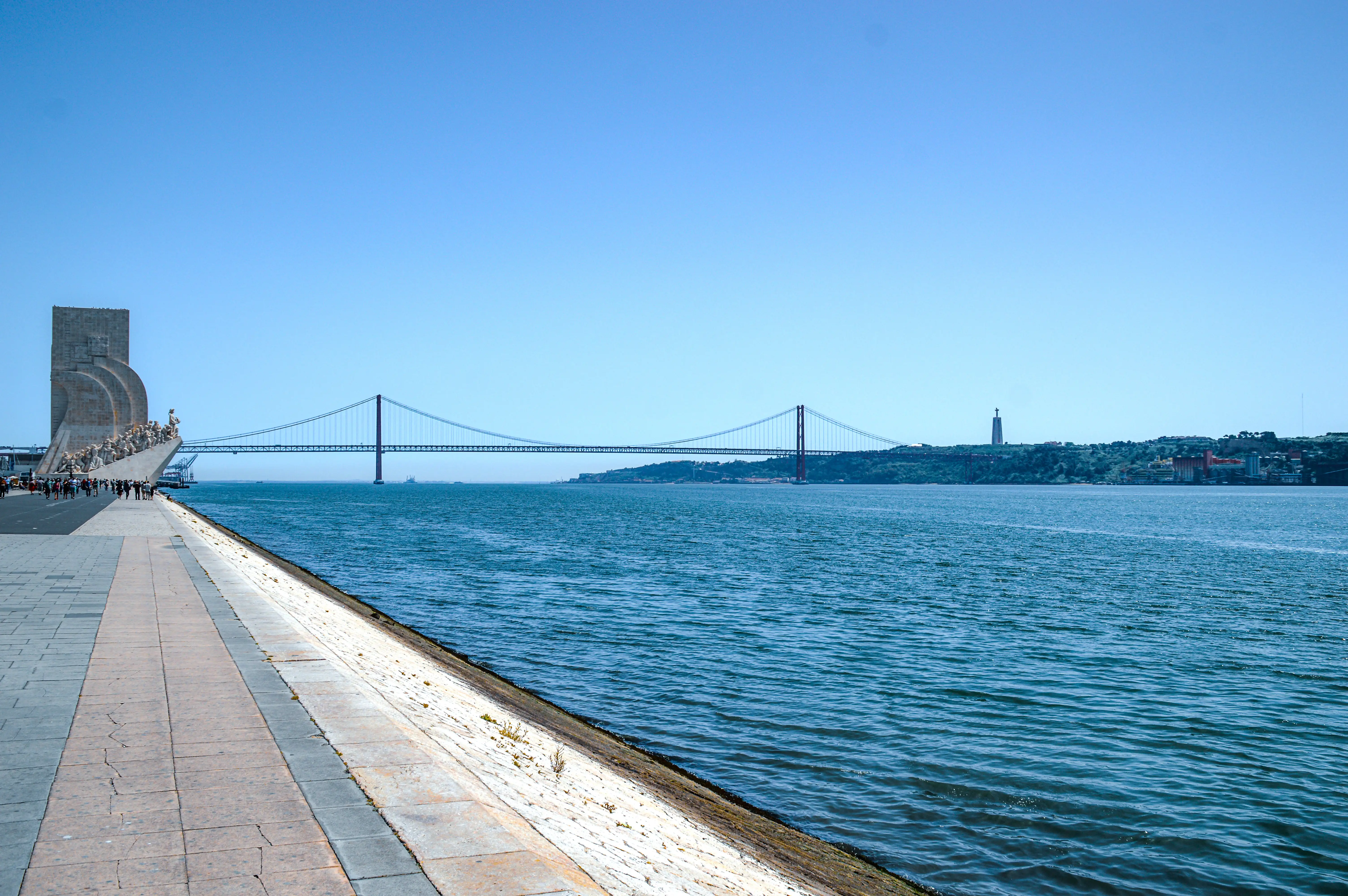 The Monument to the Discoveries on the left and the 25 de Abril Bridge spanning the Tagus River in Lisbon, Portugal, under a clear blue sky.